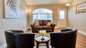 A living room with a brown leather sofa, two black leather chairs, a wooden coffee table with a vase of flowers, a large window with a view of trees, and a door with a frosted glass panel. - Cozy and Stylish Home (Boise)