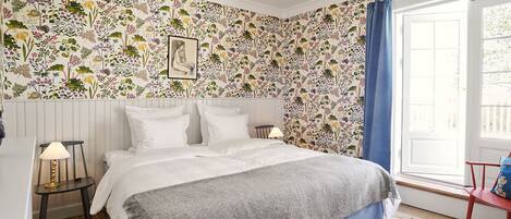 A bedroom with a floral wallpaper, a bed with white linens, a grey throw, a nightstand with a lamp, and a window with blue curtains.