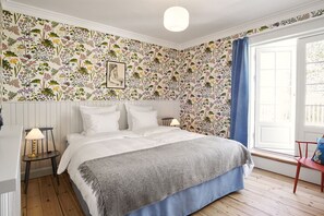 A bedroom with a floral wallpaper, a bed with white linens, a grey throw, a nightstand with a lamp, and a window with blue curtains. - Hotel Villa Strand (Hornbaek)