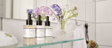 A bathroom with a glass shelf holding three bottles of CODA Paris body wash and a vase with flowers.