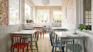 A dining area with white walls, floral wallpaper, and large windows. There are multiple tables and chairs, some with plants on them. - Hotel Villa Strand (Hornbaek)