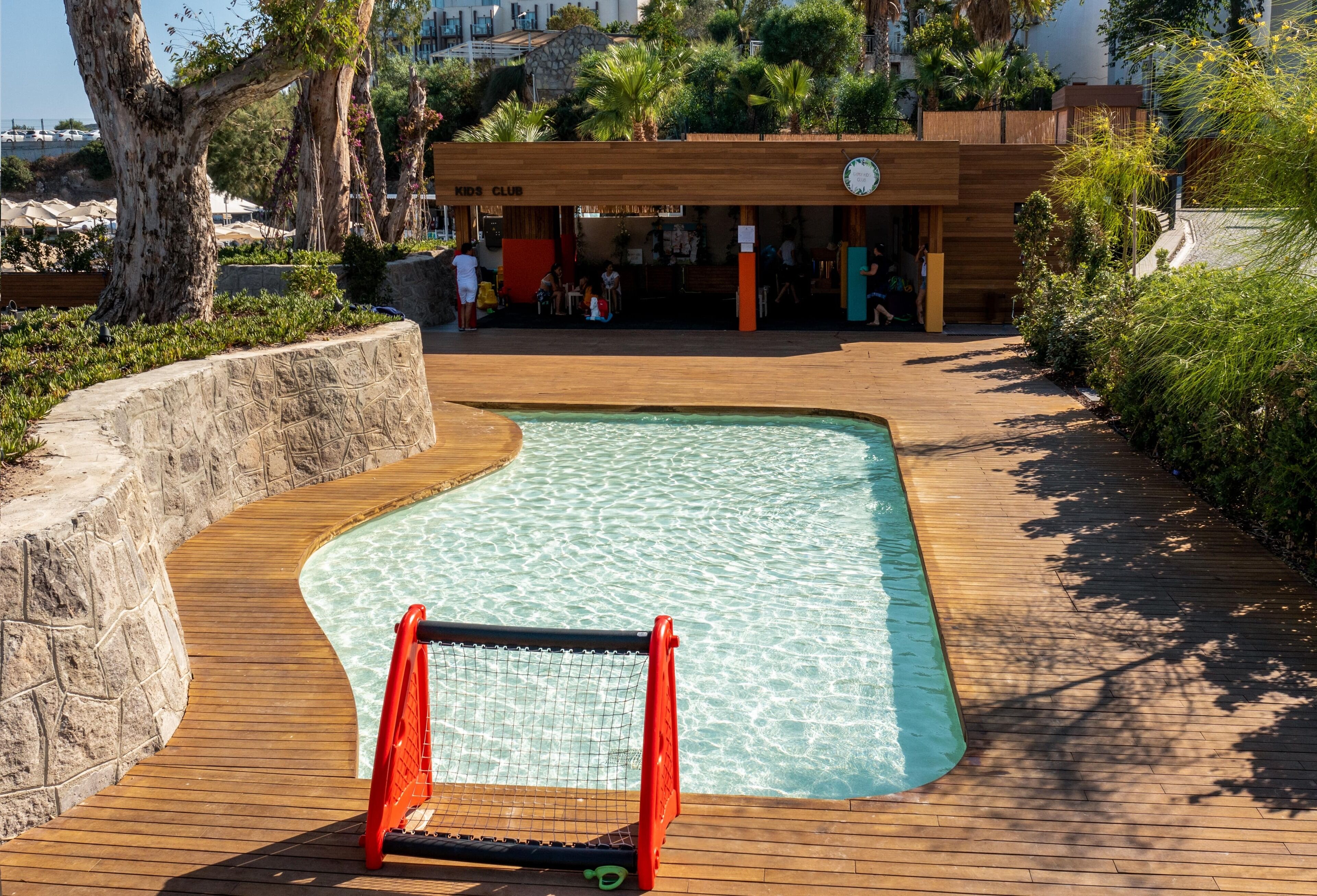 A swimming pool with a wooden deck, surrounded by greenery and a children's club building.