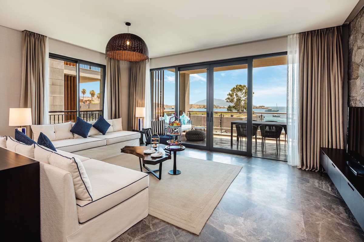 Un salon spacieux avec un canapé, une table basse et un téléviseur. Il y a un balcon avec vue sur la mer et les palmiers.