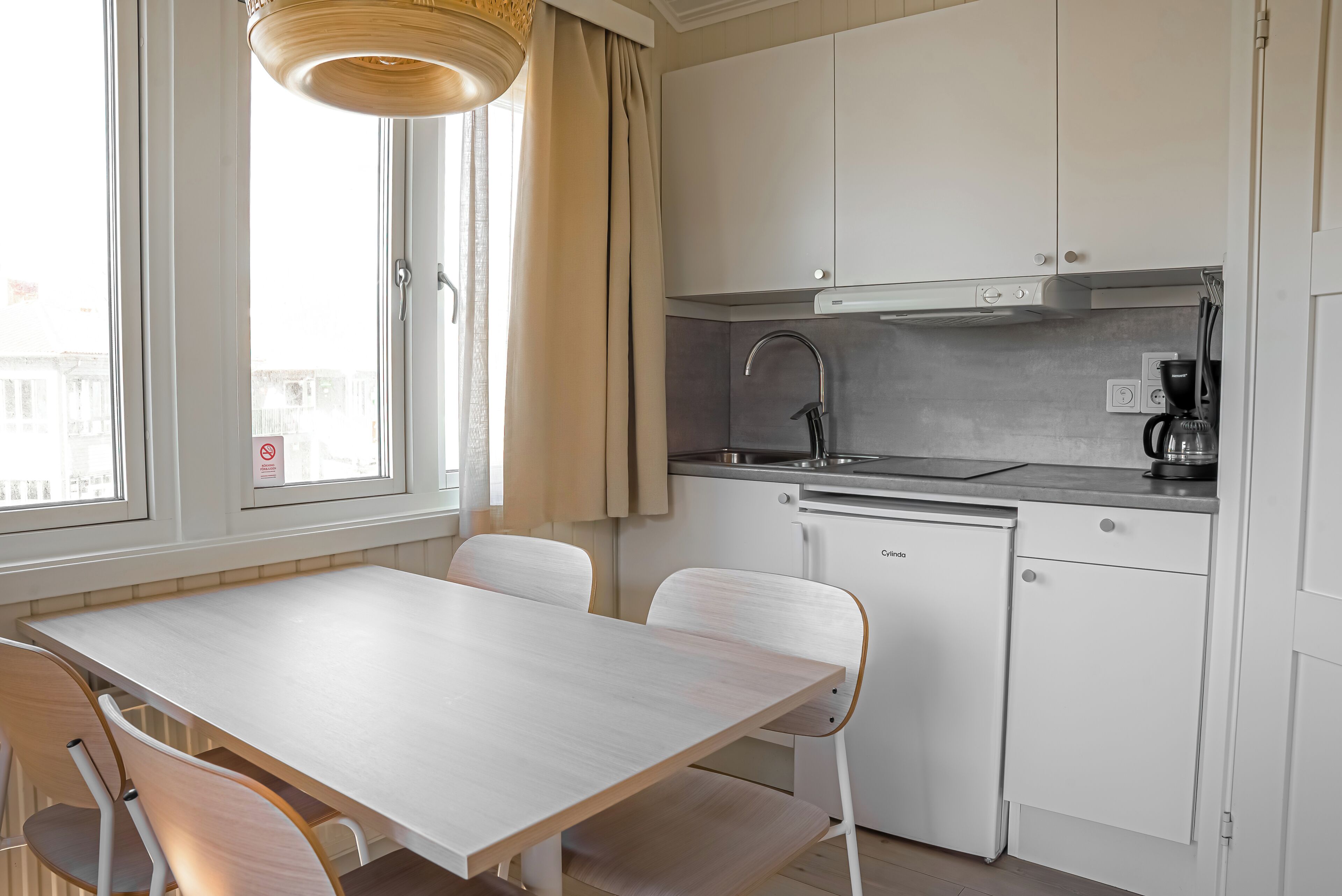 Una cocina moderna con armarios blancos, lavavajillas y cafetera. Hay una mesa de comedor con sillas y una ventana con cortinas.