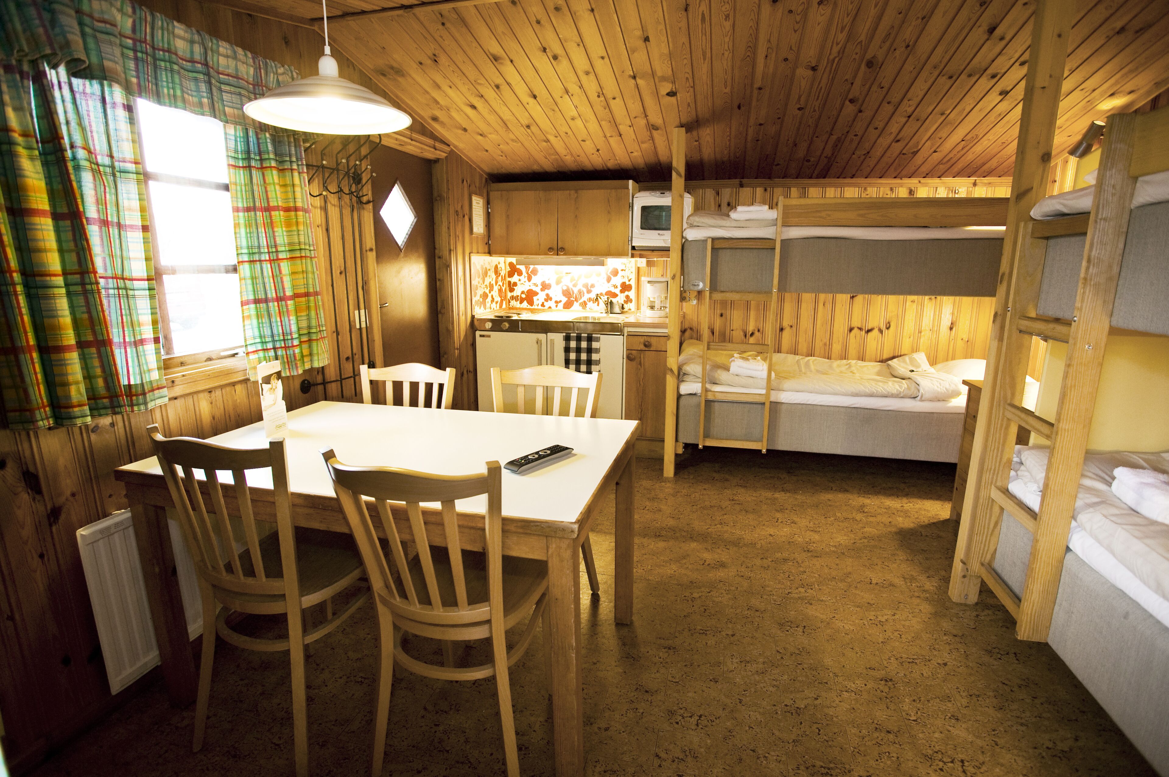 A cabin with a dining area, bunk beds, a kitchen, and a window with plaid curtains.