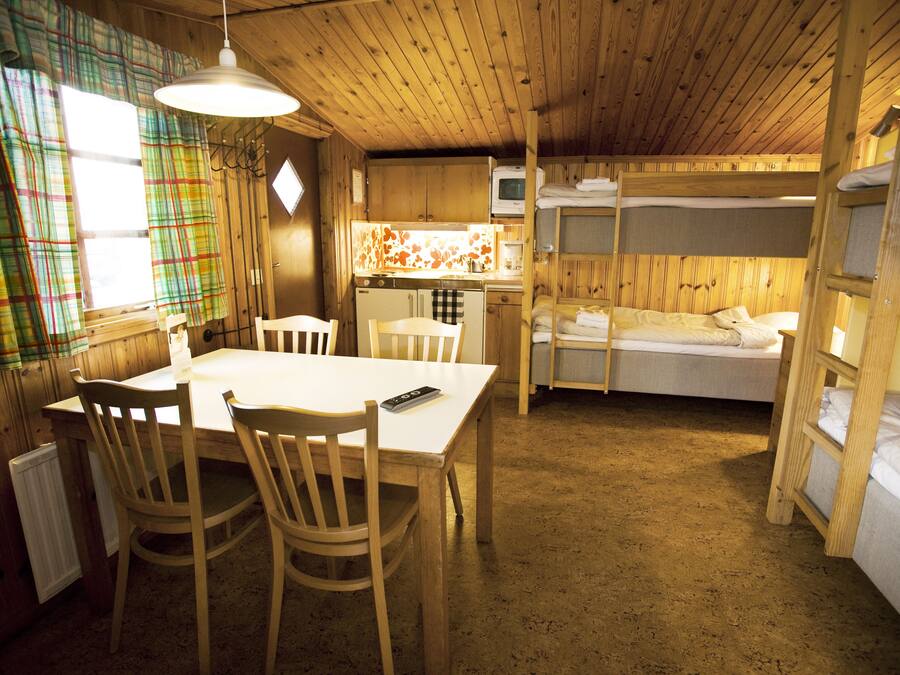 A cabin with a dining area, bunk beds, a kitchen, and a window with plaid curtains.
