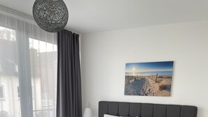 A modern bedroom with a large bed, grey upholstered bench, and a view of the outdoors through sheer curtains. - Apartment Sontje the first time OG (Laboe)