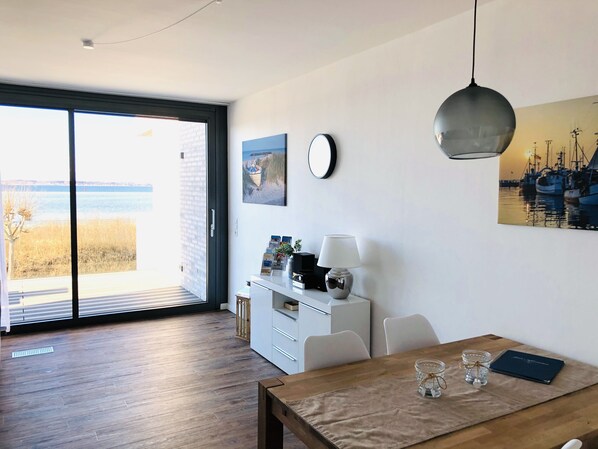 A modern living room with a wooden floor, a dining table with a placemat, a white cabinet, a lamp, and a painting of boats on the wall.