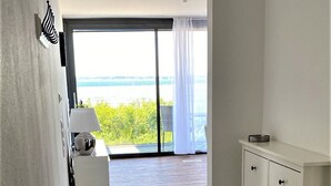 A modern room with a wooden floor, a white dresser, a small white table with a lamp, and a view of the sea through a large window. - Apartment Eltje on the 1st floor (Laboe)
