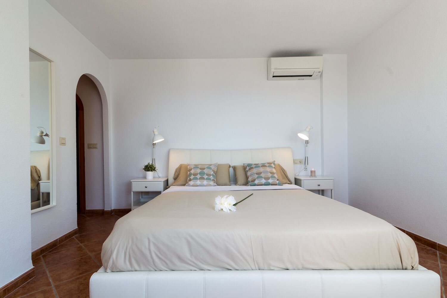 A bedroom with a large bed, two bedside tables with lamps, a wall-mounted air conditioner, and a small potted plant.