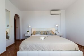 A bedroom with a large bed, two bedside tables with lamps, a wall-mounted air conditioner, and a small potted plant.