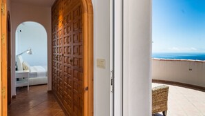 Ein Hotelzimmer mit Meerblick, einer Holztür, einem weißen Bett und einem kleinen Balkon mit Korbstuhl.