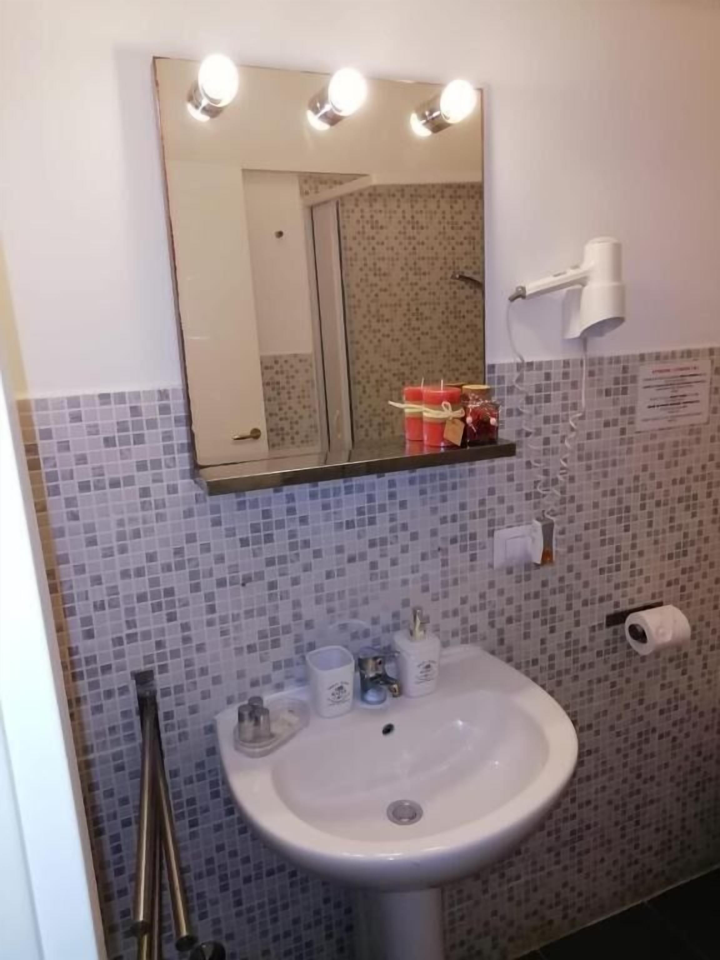 A bathroom with a white sink, a mirror with three lights, a towel rack, and a small shelf with candles.