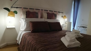 Een net opgemaakt bed met een gestreept laken, twee kussens en een set opgevouwen handdoeken ernaast. Aan weerszijden van het bed hangen twee wandlampen en erboven hangt een decoratieve orchideeënplant.