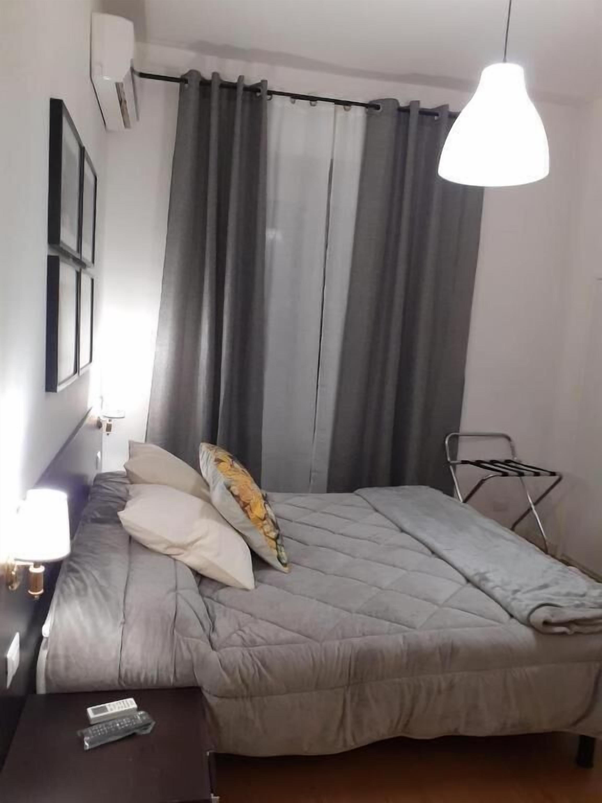 A bedroom with a bed, a bedside table with a lamp, a wall-mounted air conditioner, and a hanging light fixture.