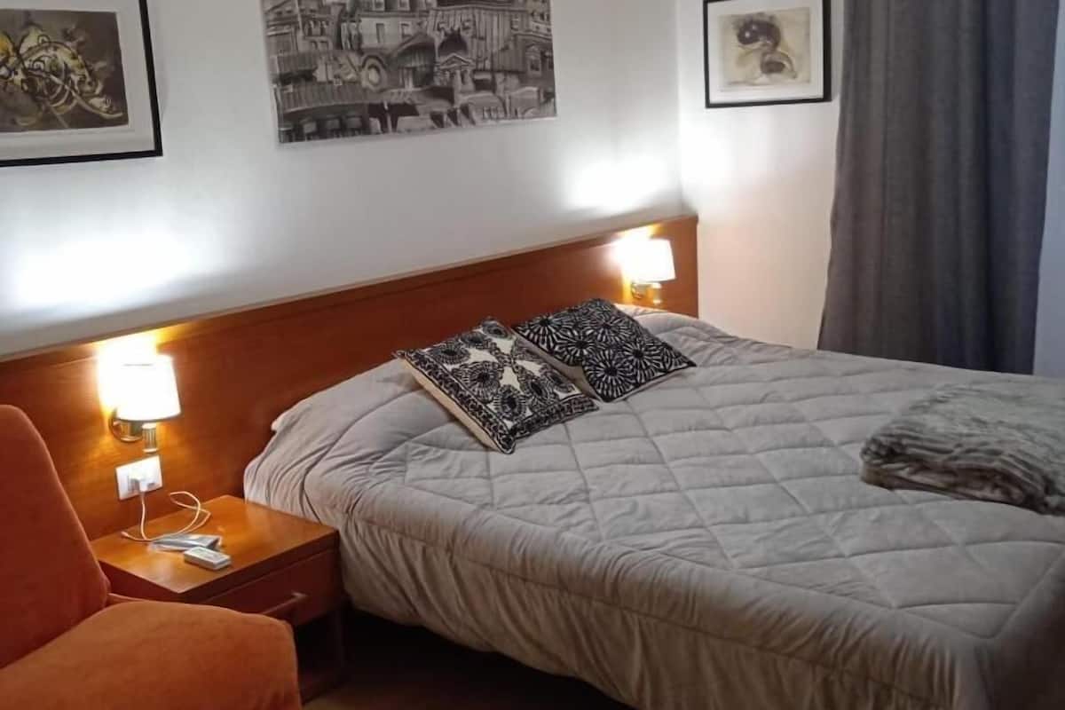 A hotel room with a bed, a nightstand, a lamp, a chair, and two framed pictures on the wall.