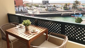 Ein Balkon mit Holztisch und Korbstühlen, mit Blick auf eine Marina mit Booten und eine Küstenstadt.