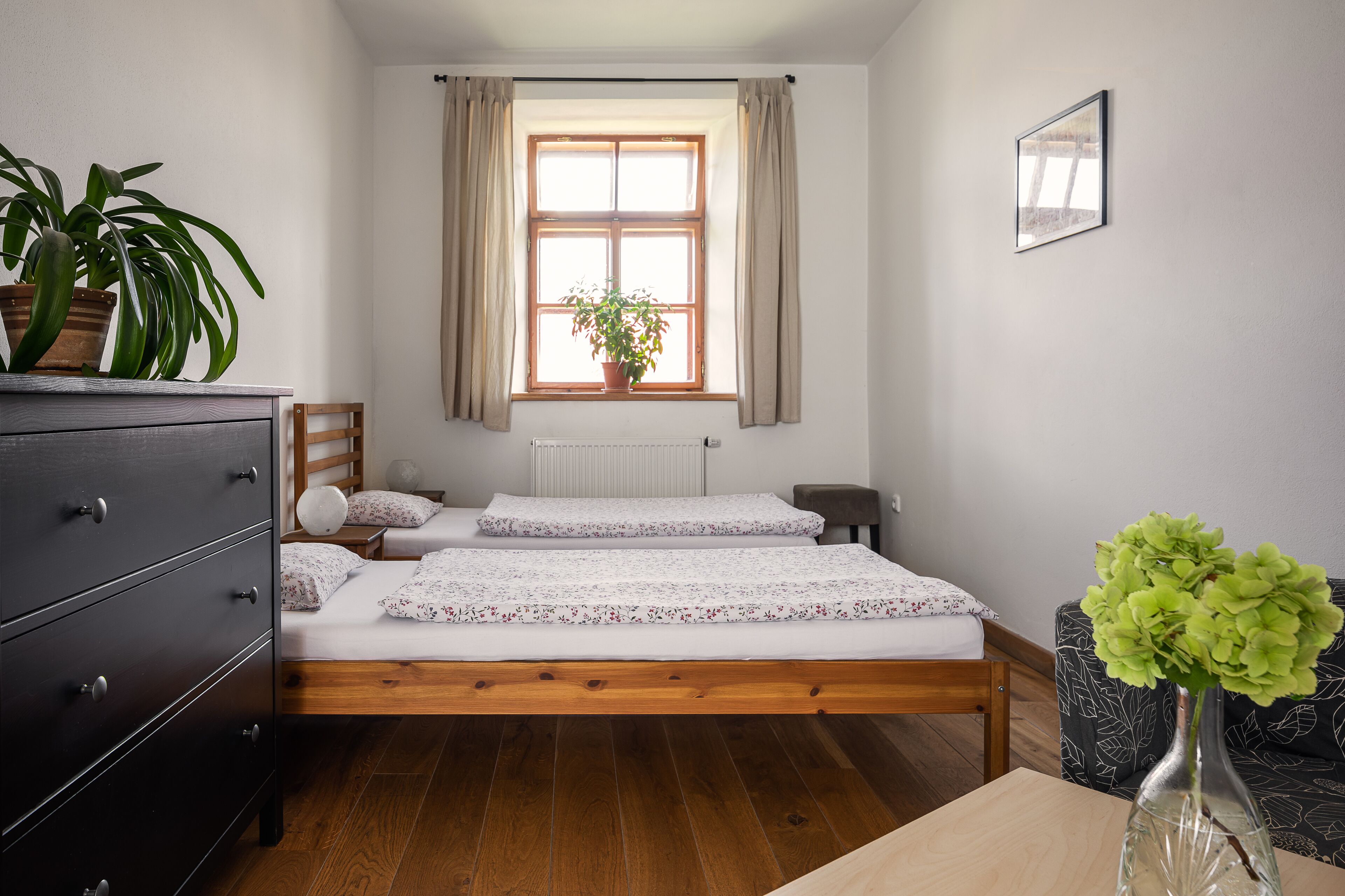 A bedroom with a wooden bed, a dresser, a window with curtains, a radiator, and a potted plant.
