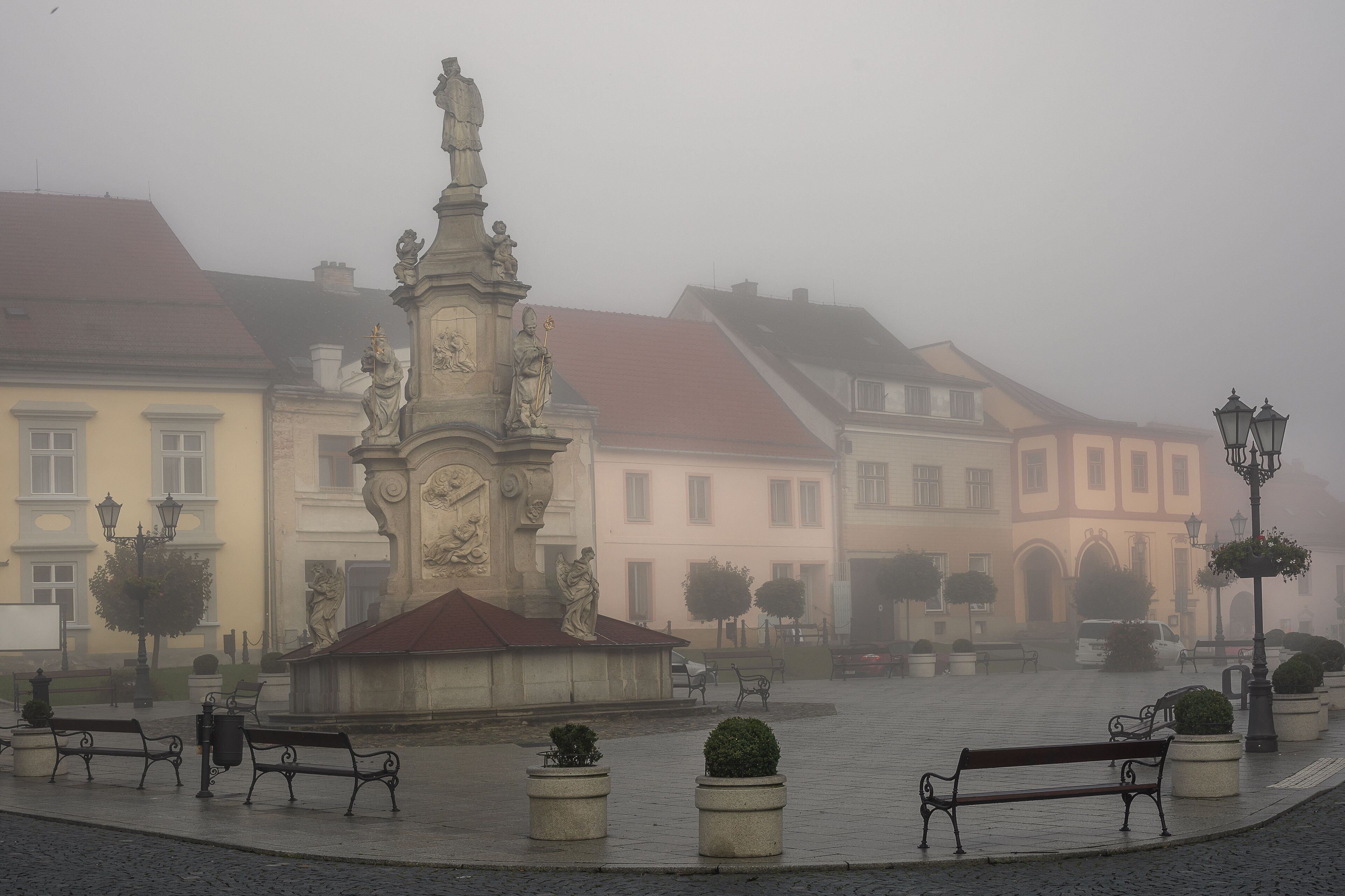 A square with benches, a statue, and buildings in the background.