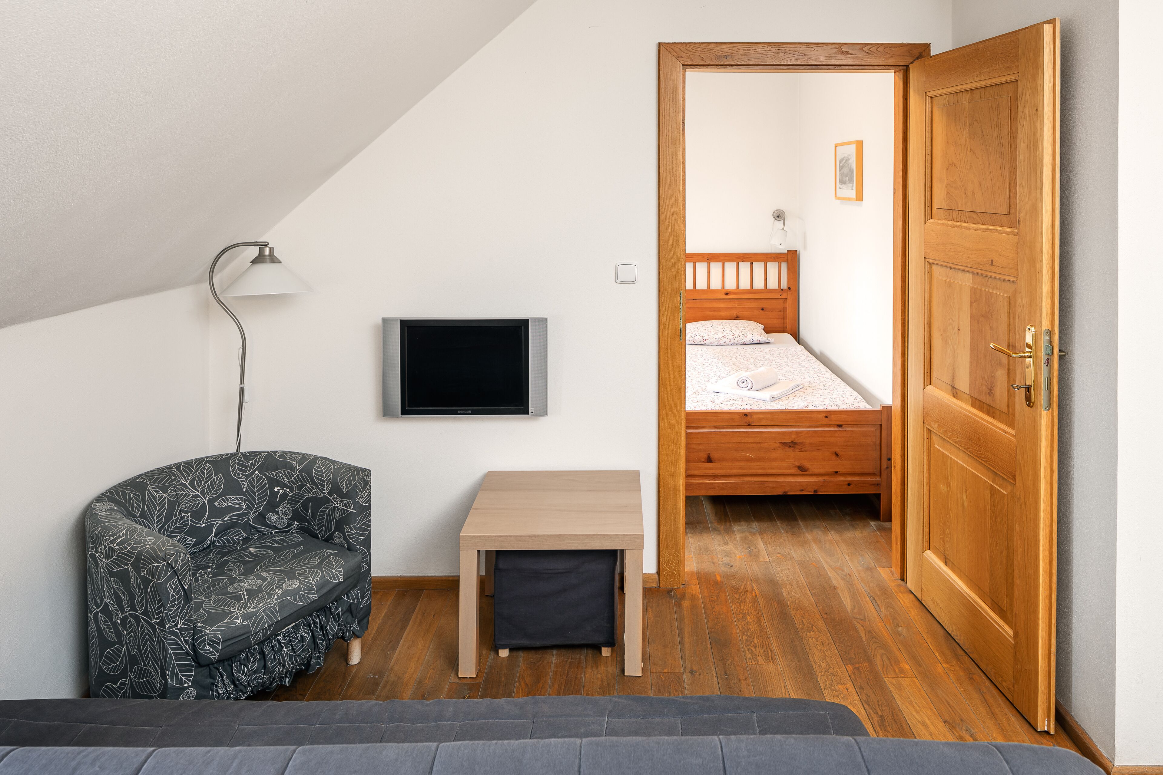 A small, well-lit room with a bed, a TV, a chair, and a wooden door.