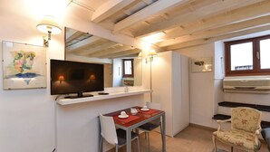 Ett litet rum med en TV, ett litet matbord med stolar, en spegel och ett fönster med persienner.