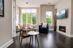 Een woonkamer met een open haard, een bank, een fauteuil en een aan de muur bevestigde televisie.