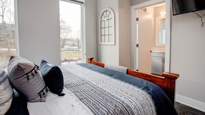Ein Schlafzimmer mit großem Fenster, einem Bett mit gemusterten Betttextilien, einem Holz-Kopfteil, einem Spiegel und einem an der Wand befestigten Fernseher.