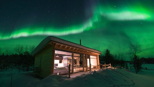 Uma cabana de madeira com fachada de vidro, rodeada por neve, sob um céu noturno com estrelas visíveis e aurora verde.