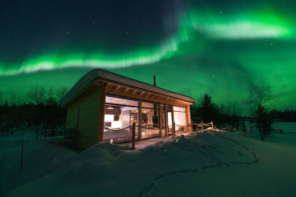 Une cabane en bois dotée d’une façade en verre, entourée de neige, sous un ciel nocturne où l’on peut voir des étoiles et des aurores boréales vertes.