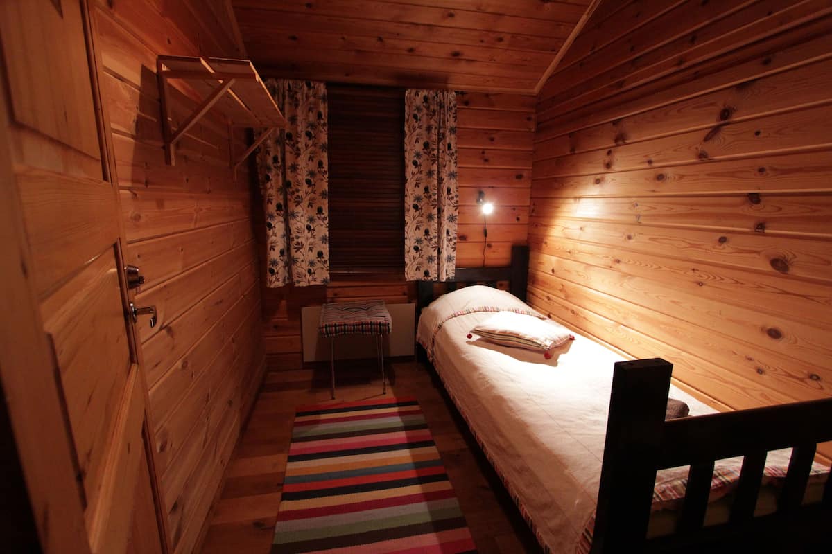 Une petite pièce chaleureuse aux murs en bois et dotée d’un seul lit. On y trouve un banc et une petite table avec une lampe.