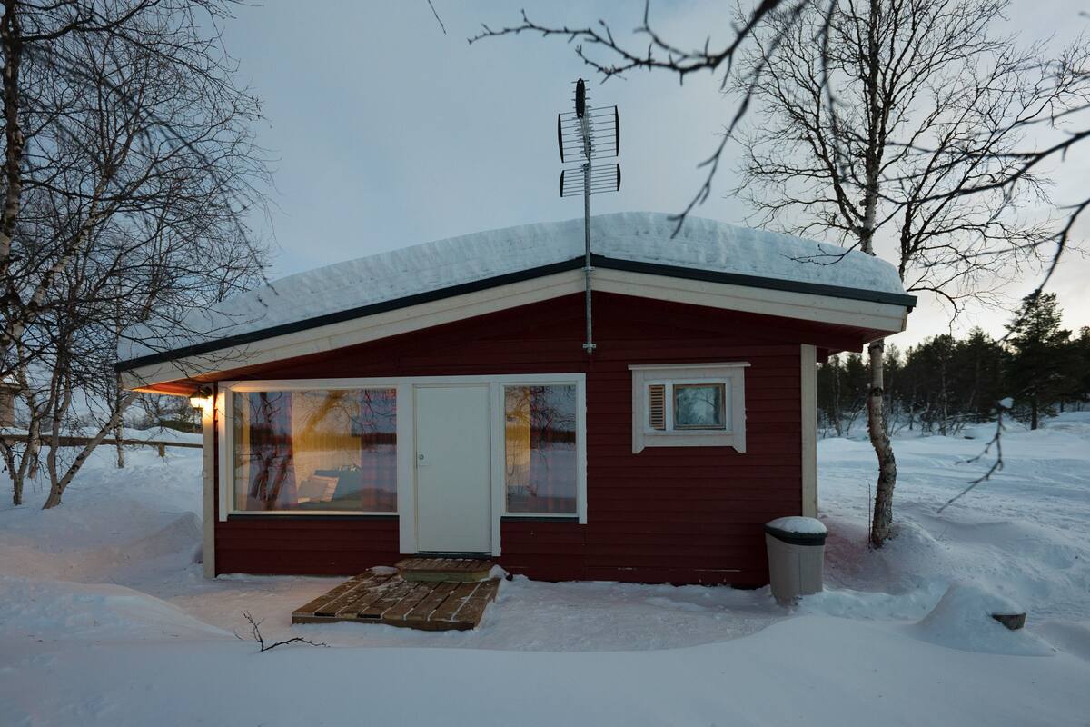 Une petite cabane rouge, avec un toit enneigé, une grande fenêtre et une porte.