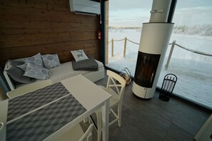 A cozy room with a wooden wall, a sofa with patterned cushions, a table, and a large window overlooking a snowy landscape. - Giellajohka (Inari)