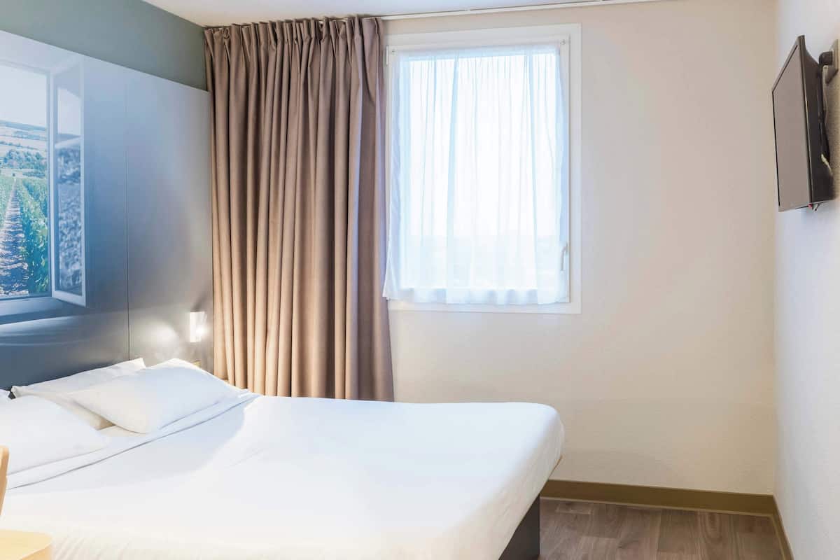A hotel room with a bed, a window with curtains, a wall-mounted TV, and a view of greenery outside.