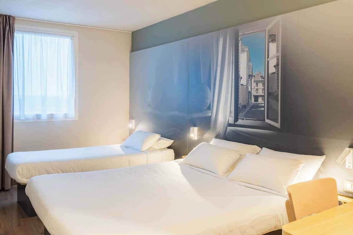A hotel room with two beds, a window with curtains, a painting of a cityscape, and a wooden table.