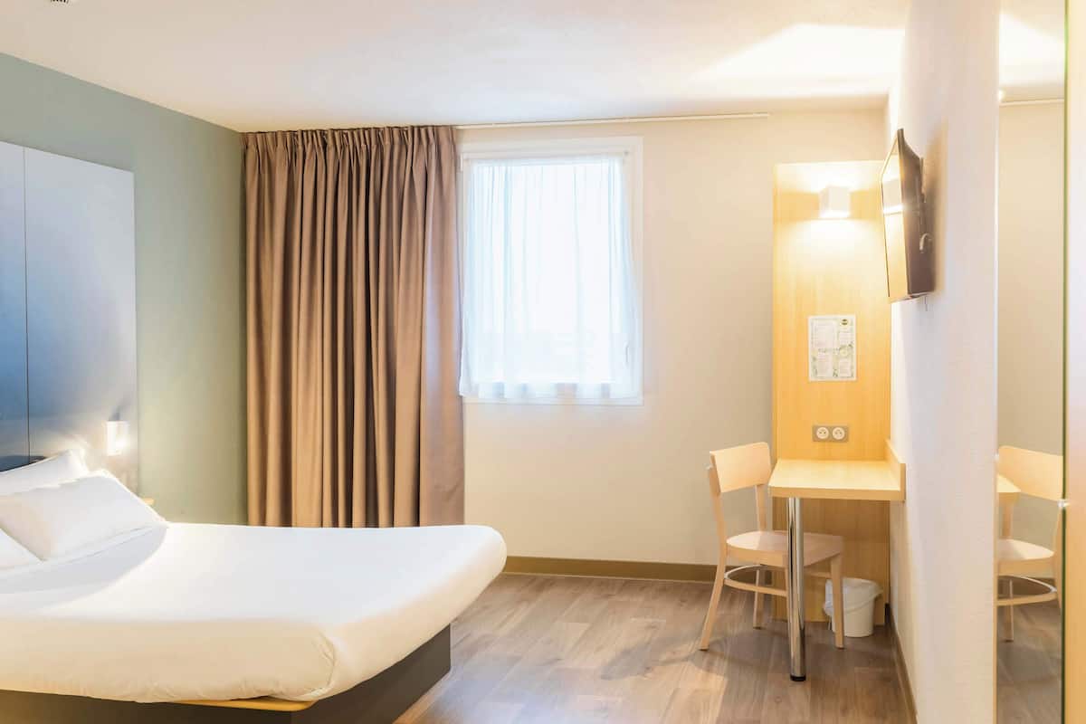 A hotel room with a bed, a desk, two chairs, a window with curtains, a wall-mounted light, and a trash bin.