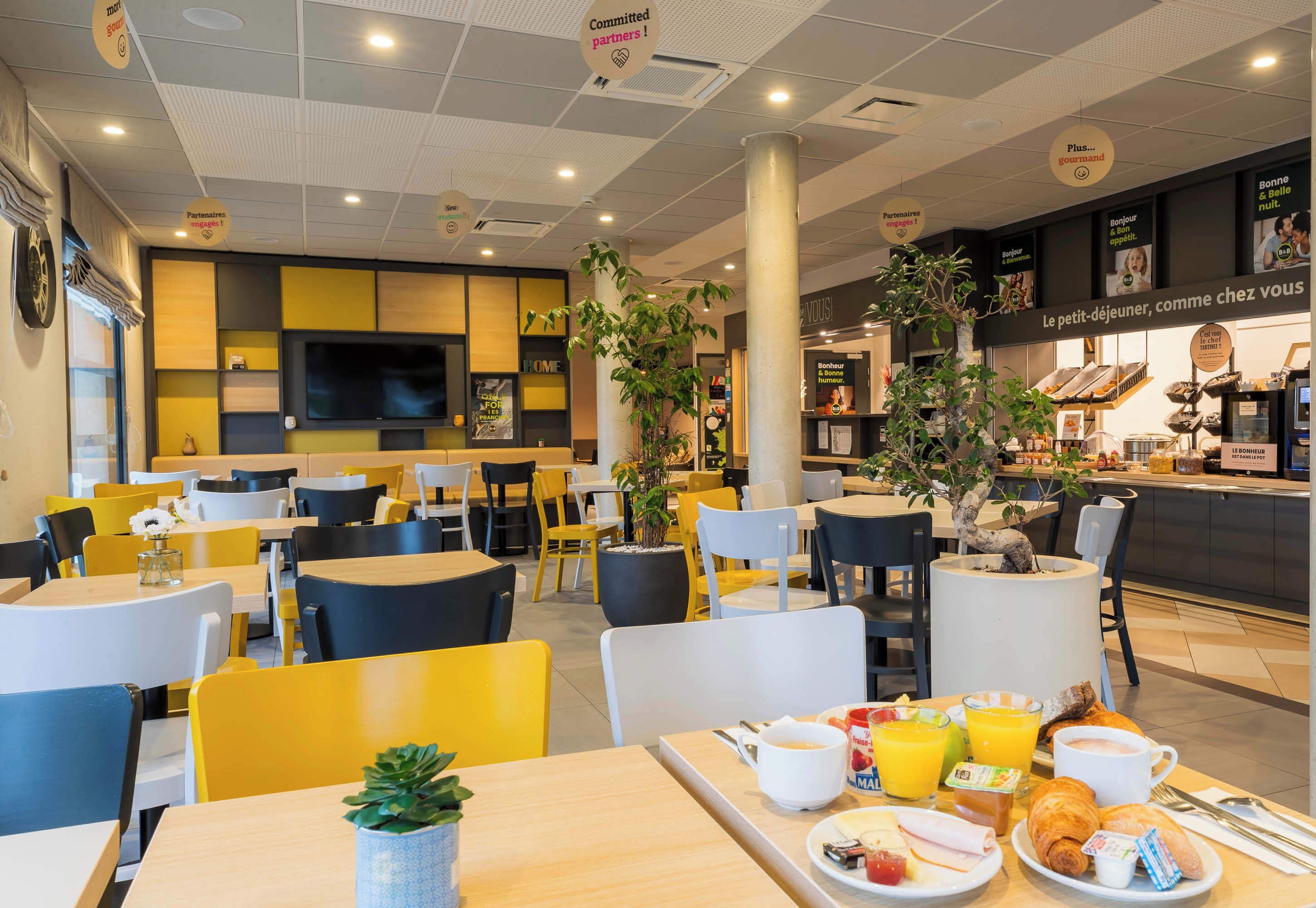 Een modern café met gele en witte stoelen, een ontbijtbuffet en een televisiescherm aan de muur.