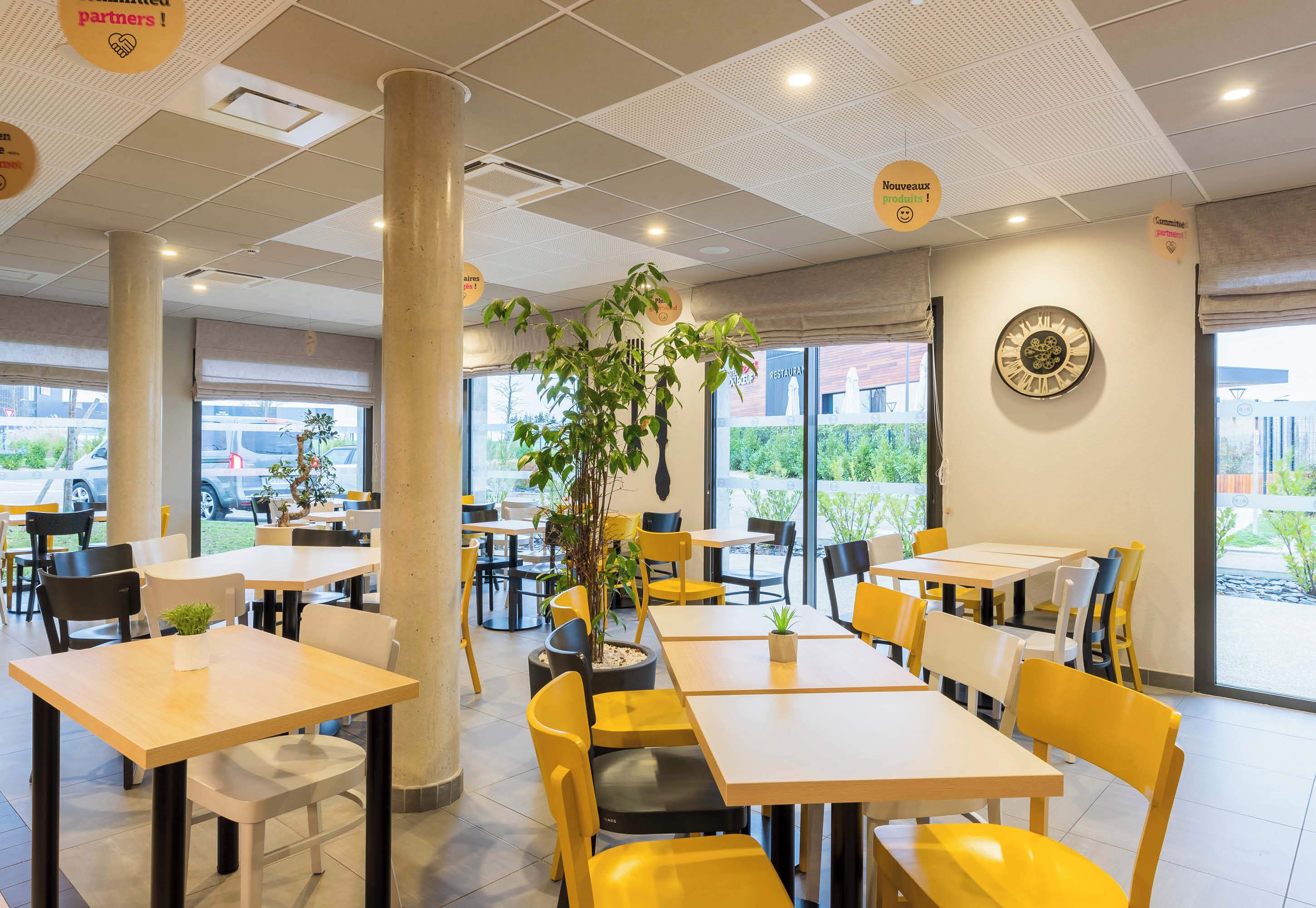 A modern indoor dining area with tables and chairs, large windows, and a decorative wall clock.