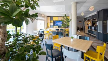 A modern restaurant with a wooden table and yellow chairs, a large pillar, and a TV screen on the wall.