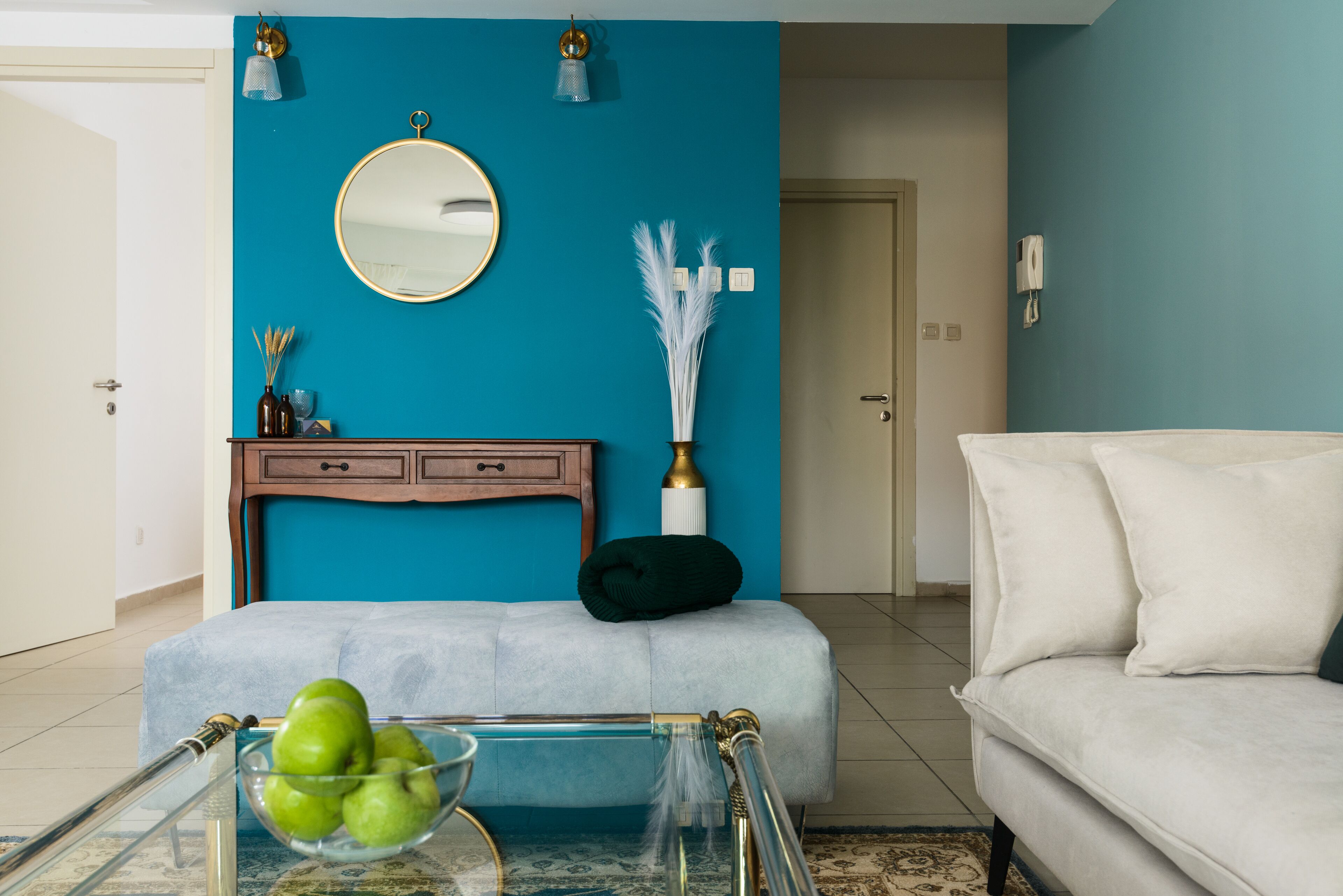 Een woonkamer met teal muren, een ronde spiegel, een witte bank en een glazen salontafel met appels.