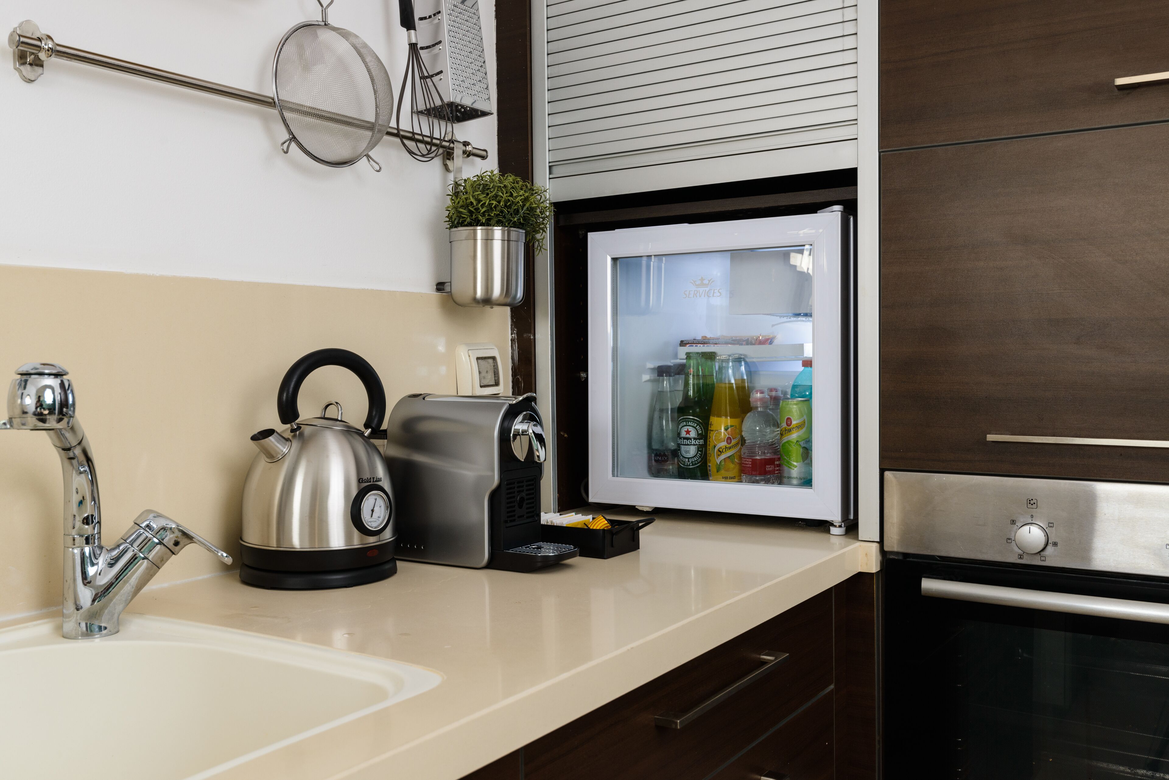 A kitchen with a stainless steel kettle, a coffee maker, a refrigerator stocked with beverages, and a sink.