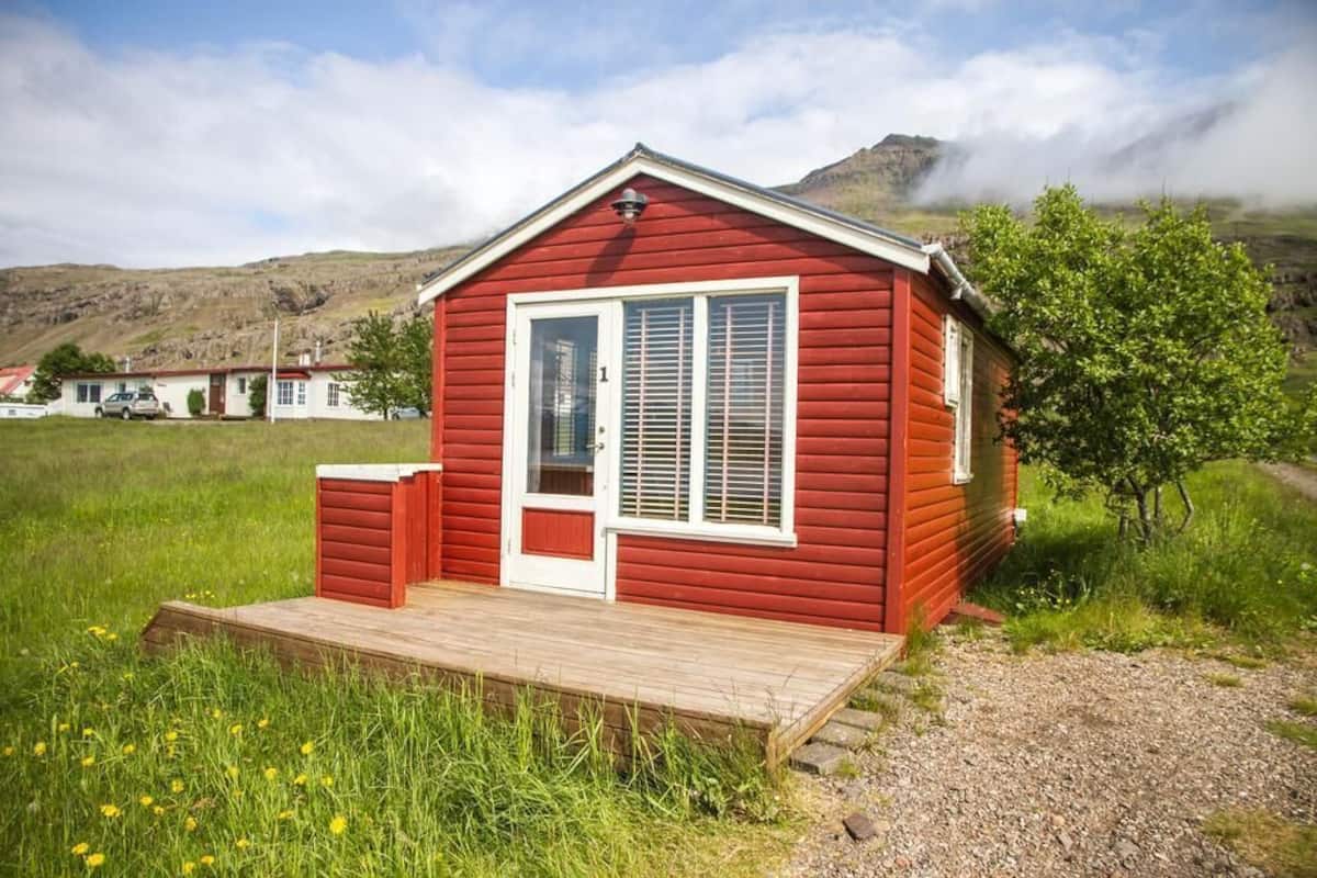 A red wooden cabin with a white door and window shutters, a wooden deck, and a grassy field with a mountain in the background.