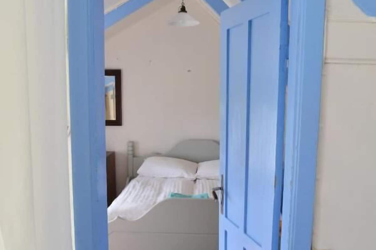 A narrow hallway with a wooden floor leading to a bedroom with a bed and a single hanging light fixture.
