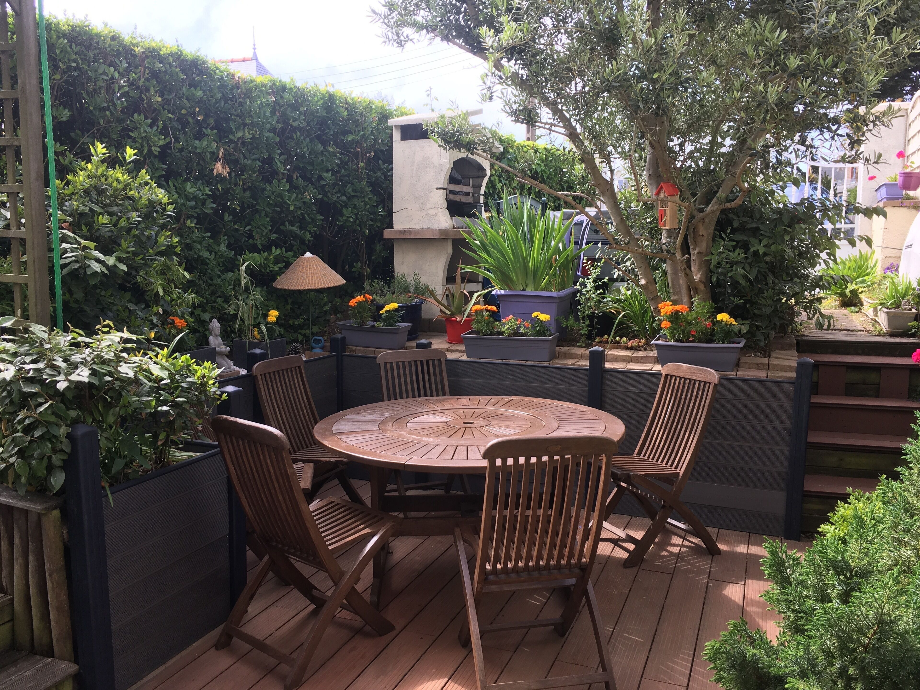 Un patio con una mesa redonda de madera y cuatro sillas, rodeado de macetas con plantas y un seto.
