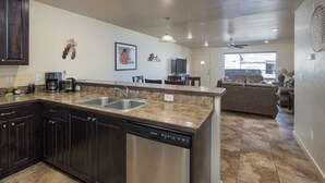 Una cocina con encimeras de granito, electrodomésticos de acero inoxidable y armarios de madera oscura. Hay una zona de comedor con un sofá y un ventilador de techo.