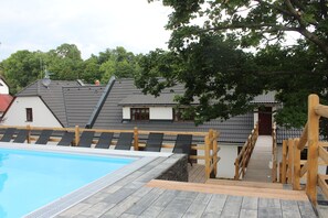 A wooden deck leading to a swimming pool with lounge chairs, surrounded by houses with sloped roofs and a large tree. - Relax Penzion Schonwald (Budišov nad Budišovkou)