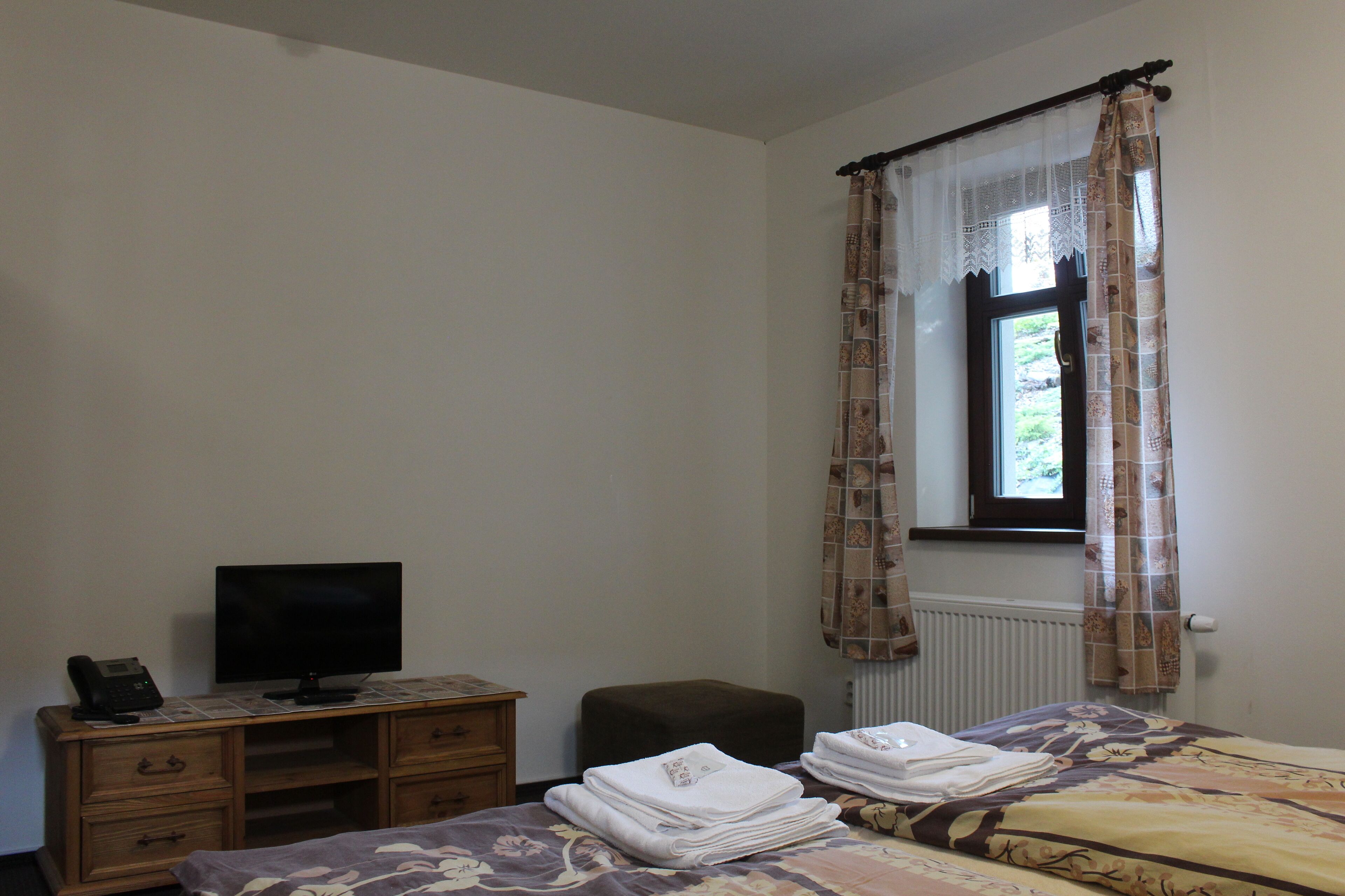 Un dormitorio con cama, televisor sobre una cómoda de madera, ventana con cortinas y radiador.