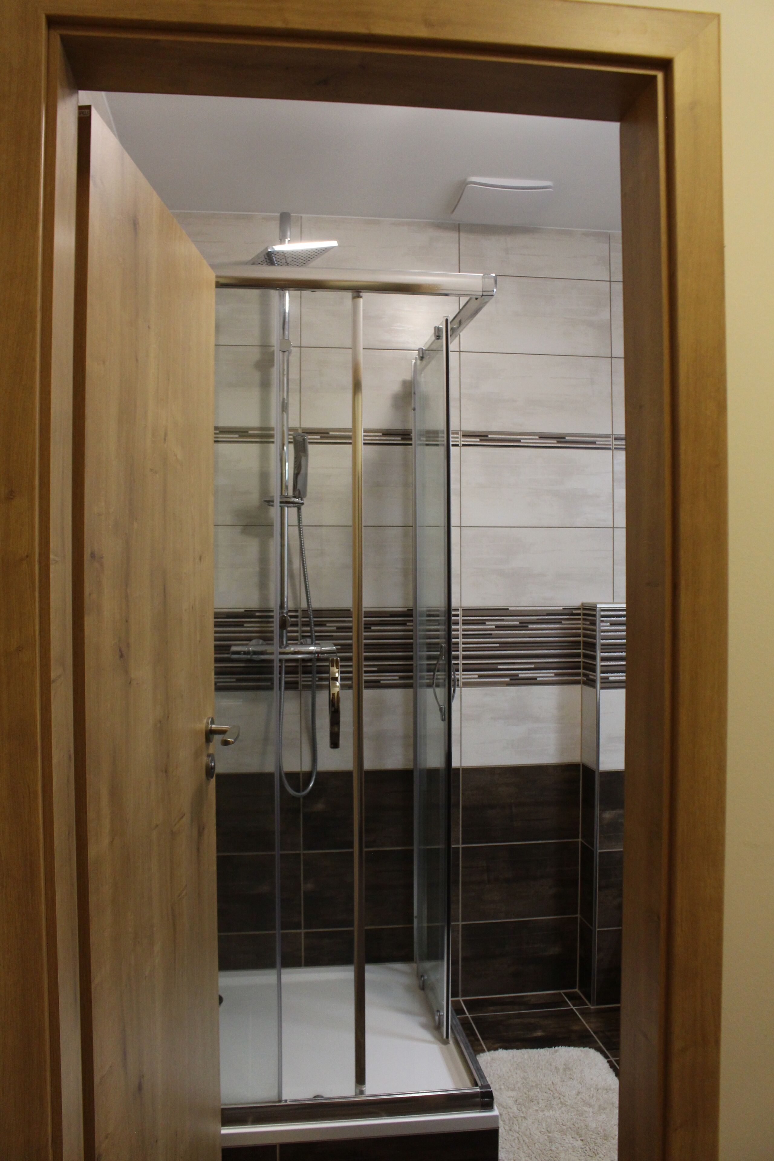 A glass-enclosed shower with a chrome showerhead and control panel, a white tiled floor, and a wooden door frame.