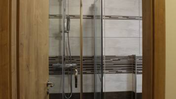 A glass-enclosed shower with a chrome showerhead and control panel, a white tiled floor, and a wooden door frame.