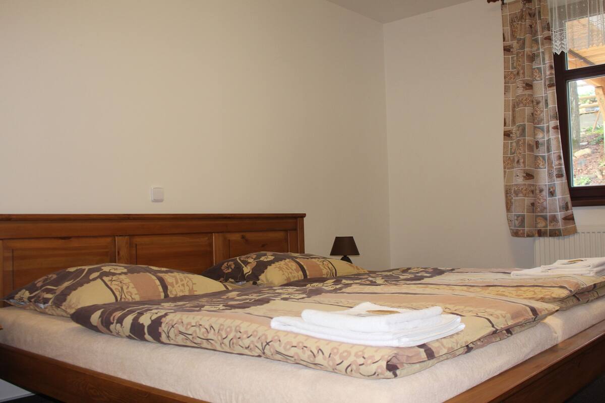 Ein ordentlich bezogenes Bett mit einer blumigen Tagesdecke und Kissen, ein Holzkopfstück und ein Fenster mit gemusterten Vorhängen.