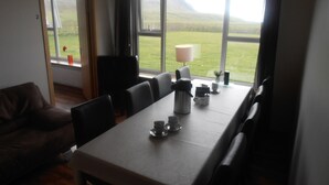 A dining area with a table set for four, chairs, a coffee pot, cups, and a lamp. A window offers a view of a green field and mountains. - Giljur Guesthouse (Vik I Myrdal)